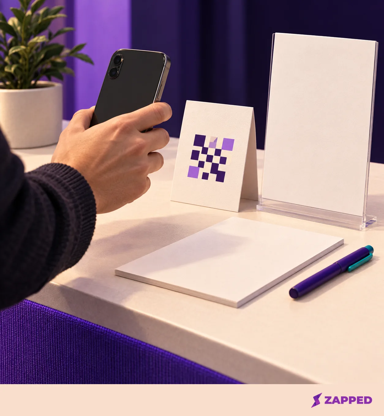 A hand aims a phone with its screen turned away at an abstract QR proof beside blank booth materials.